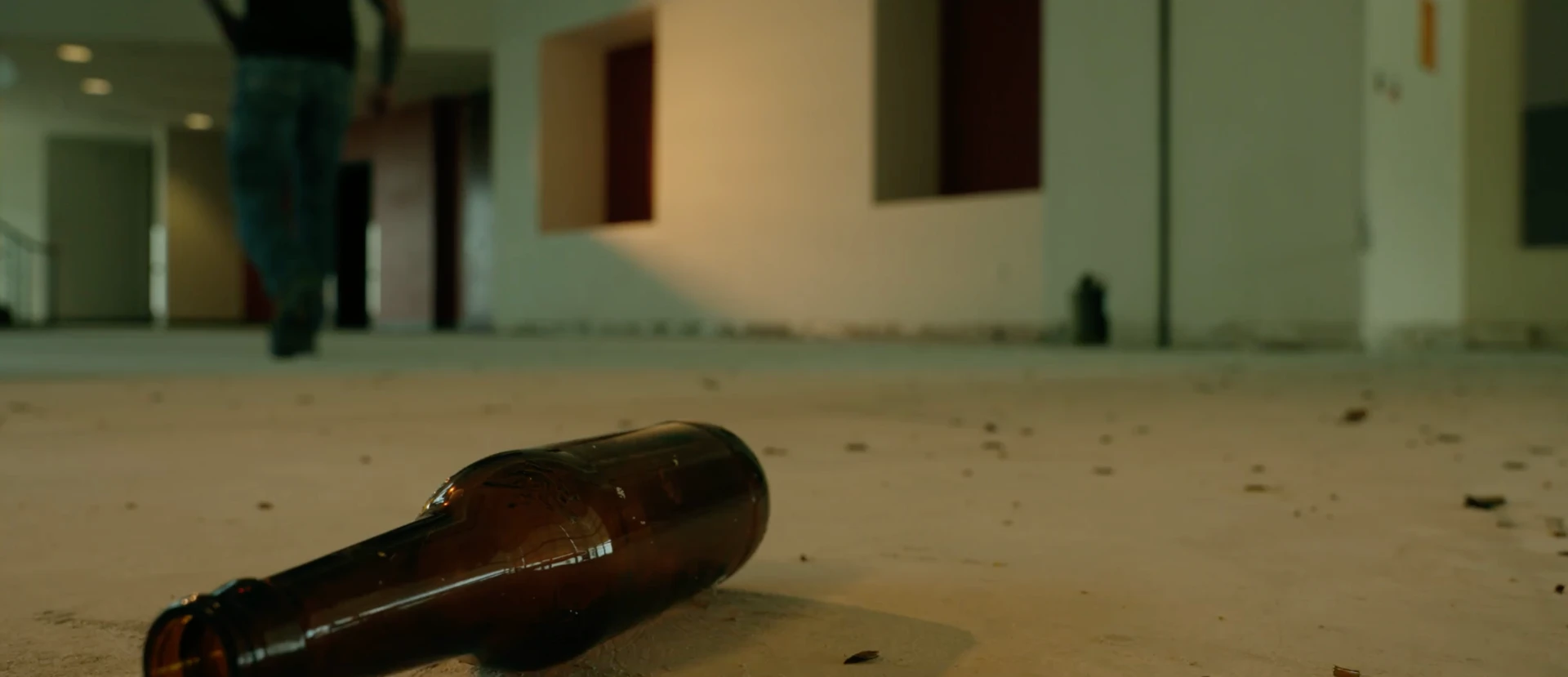An empty beer bottle on the floor as a figure walks away down an empty corridor